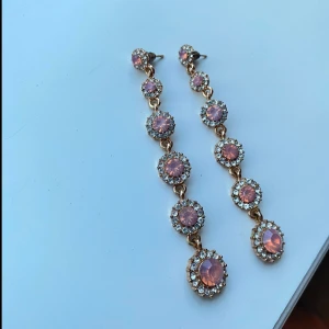 Långa örhängen med rosa stenar - Eleganta långa örhängen med flera rosa stenar omgivna av glittrande kristaller. Perfekta för att ge en glamorös touch till din outfit. De har en gyllene bas och hängande design.
