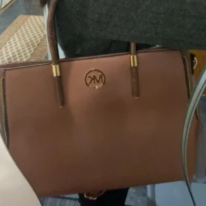 Brun axelväska från Michael Kors - Snygg brun axelväska från Michael Kors med klassisk design. Väskan har två handtag och en avtagbar axelrem. Den är tillverkad i skinn och har en dragkedja på baksidan för extra förvaring. Perfekt för att bära dina dagliga nödvändigheter med stil.