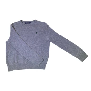 Grå tröja från Polo Ralph Lauren 100% merino ull - Säljer en stilren grå tröja från Polo Ralph Lauren med rund hals och det klassiska logotypbroderiet i grönt på bröstet. Tröjan har långa ärmar och ribbade muddar vid ärmslut och nederkant. 100% merino ull