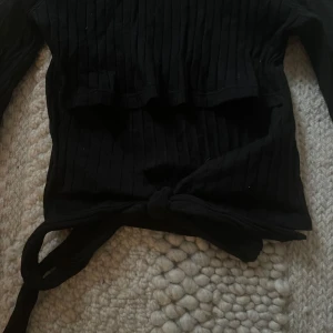 Svart ribbad tröja från NA-KD - Säljer en stilren svart ribbad tröja från NA-KD. Tröjan har långa ärmar och en knytning baktill som ger en snygg detalj. 