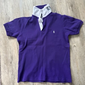 Lila pikétröja från Ralph Lauren - Säljer en snygg lila pikétröja från Ralph Lauren med vit krage och knappar. Tröjan har korta ärmar och den klassiska loggan broderad på bröstet. Perfekt för en stilren look!  Passar mer som s än M!