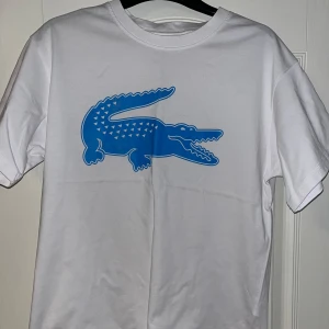 Lacoste Vit t-shirt  - Säljer en vit t-shirt från Lacoste med en stor blå krokodil på framsidan. T-shirten har en klassisk passform och är kortärmad. Perfekt för en avslappnad stil.