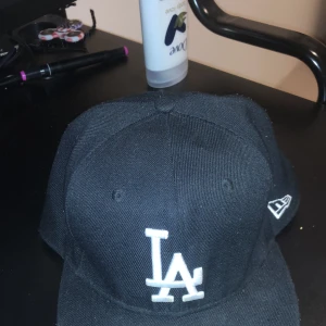 Svart keps från New Era - Snygg svart keps från New Era med LA-logga i vitt framtill. Kepsen är en 59FIFTY-modell med en rak skärm och broderad logga på sidan. Perfekt för en casual look.