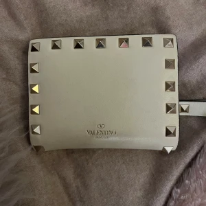 Vit plånbok från Valentino - Snygg kräm vit plånbok från Valentino med nitar i guldton runt kanterna. Plånboken har flera kortfack och en knappstängning. Perfekt för att hålla ordning på dina kort och kontanter med stil. Jag köpte den i somras för 2000 på kindal i japan. Syns lite användning.