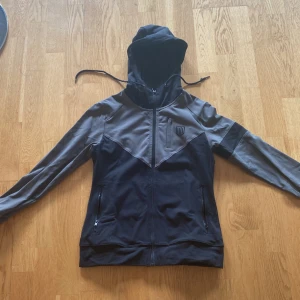 Iris V Hoodie från Maya Delorez  - Säljer en snygg och praktisk svart och grå vindjacka från Maya Delorez. Jackan har en dragkedja framtill och en huva med dragsko. Perfekt för blåsiga dagar och designad i Sverige. Den har en liten logga på bröstet och en ficka med dragkedja. Passar perfekt för vår och höst!