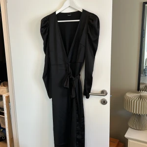 Svart omlottklänning från Gina Tricot - Säljer en elegant svart omlottklänning från Gina Tricot. Klänningen har långa ärmar med en puffig design och ett knytband i midjan för en smickrande passform. Perfekt för festliga tillfällen eller en kväll ute. 🖤