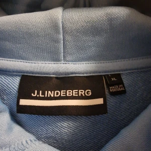 Ljusblå hoodie från J.Lindeberg - Säljer en superfin ljusblå hoodie från J.Lindeberg i samarbete med Nelly Korda. Den är i storlek XL och har en klassisk känguruficka framtill. Hoodien har en skön passform och är perfekt för både vardag och chill. Den har vita snören vid huvan och texten J.LINDEBERG' broderad på bröstet. Perfekt för höst och vår!