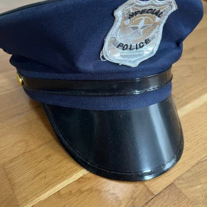 Mörkblå polisliknande hatt - Säljer en mörkblå polisliknande hatt med en blank svart skärm och ett silverfärgat märke framtill. Perfekt för maskerad eller temafest! Hatten har en klassisk stil och är i bra skick.