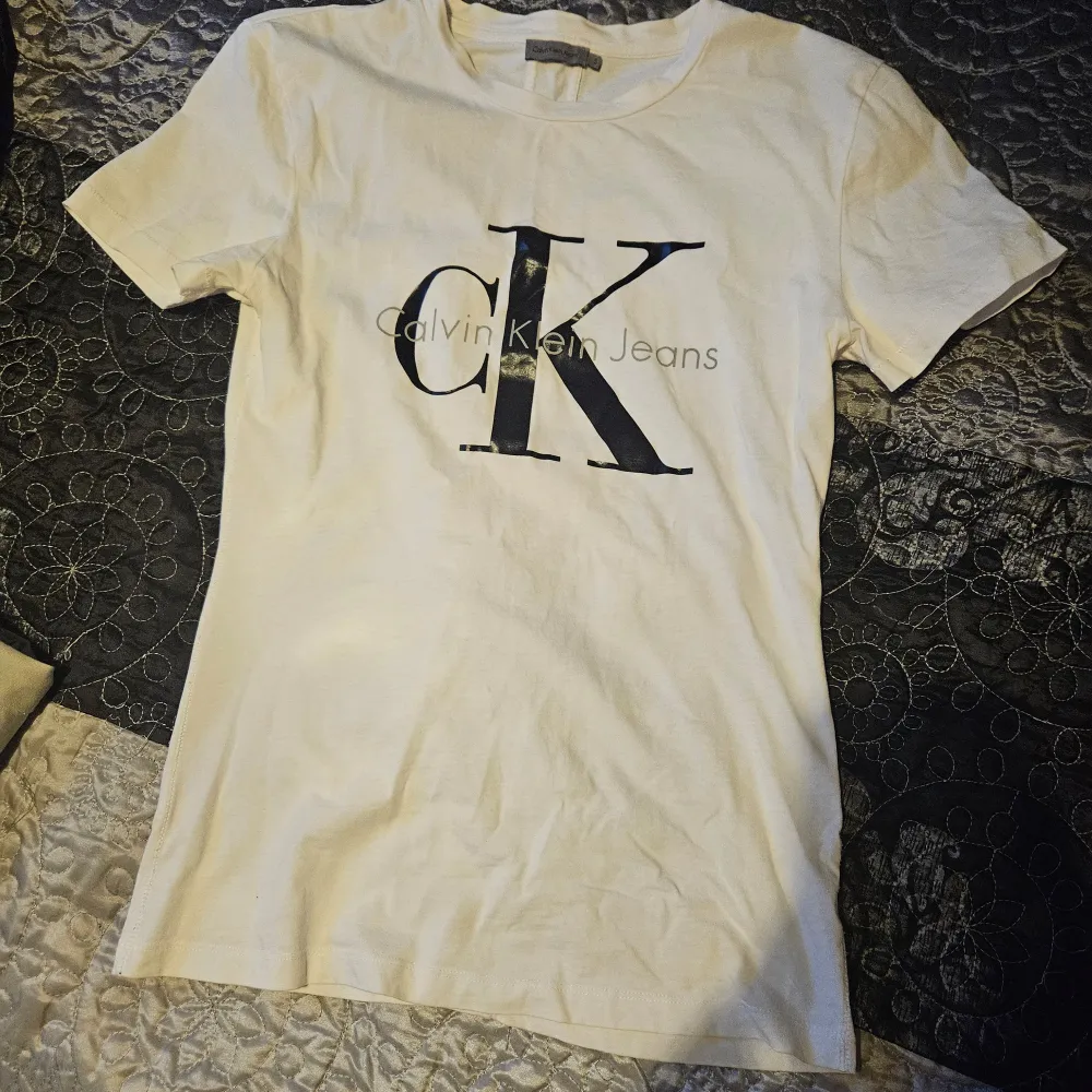 Säljer en stilren vit t-shirt från Calvin Klein Jeans. Den har en stor CK-logga på framsidan i svart. T-shirten är kortärmad och gjord i mjukt bomullsmaterial. Perfekt för en avslappnad och trendig look!. T-shirts.