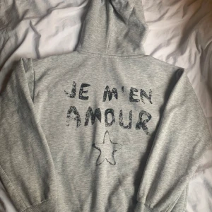 Grå hoodie med text - Säljer en supermysig grå hoodie med texten 'JE M'EN AMOUR' och en stjärna tryckt på både fram- och baksidan. Perfekt för en chill dag eller när du vill ha något bekvämt och stilrent. Den har långa ärmar och en klassisk huva. Passar perfekt till jeans eller mjukisbyxor! Den är lite nopprig därav priset