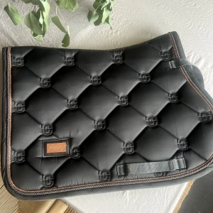 Svart quiltad sadelpad från Equestrian Stockholm - Säljer en snygg och stilren svart quiltad sadelpad från Equestrian Stockholm. Den har en elegant design med bruna detaljer och är perfekt för ponny. Padden är i nyskick och har en lyxig känsla med sitt mjuka material och fina sömmar. den har bara använd en gång!