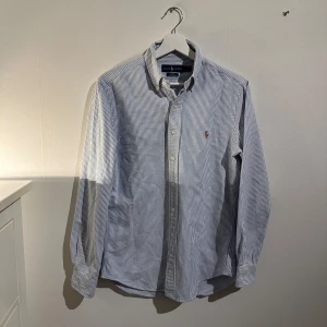 Randig skjorta från Ralph Lauren - Säljer en snygg och stilren skjorta från Ralph Lauren i ny skick skjortan är storlek S. Den är vit och blå med ett klassiskt randigt mönster. Skjortan är slim fit och gjord i stretchigt oxford-material, vilket gör den både bekväm och elegant. Perfekt för både vardag och fest! 💙