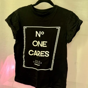 Svart t-shirt med tryck - Snygg svart t-shirt med ett djärvt vitt tryck 'No One Cares'. Perfekt för en avslappnad stil med en touch av attityd. Kortärmad och i en bekväm passform.