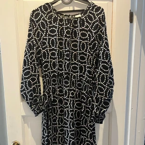 Svart och vit mönstrad klänning - Snygg svart klänning med vitt prickmönster. Den har långa ärmar och en smickrande passform. Perfekt för en stilren look. Klänningen har resor i slutet av ärmarna samt i midjan. 