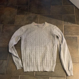 Grå stickad tröja - Säljer en stilren grå stickad tröja med kabelstickat mönster. Tröjan har långa ärmar och rund halsringning, perfekt för kyligare dagar. Passar bra till både jeans och kjol.