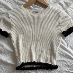 Beige ribbad topp från Zara - Säljer en snygg beige ribbad topp från Zara med svarta volangdetaljer vid ärmslut och nederkant. Toppen har korta ärmar och en cropped. knappast använd. skriv om ni har frågor.  