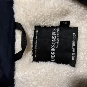 Mörkblå parkajacka från Didriksons st 150 - Säljer en mörkblå parkajacka från Didriksons med fuskpäls på huvan och fleecefoder. Jackan har långa ärmar, dragkedja och knappar framtill samt flera praktiska fickor. Perfekt för kalla dagar.