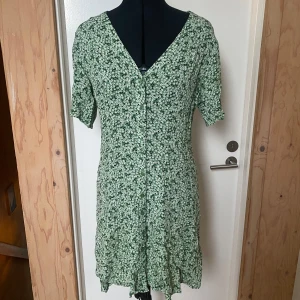 Grön blommig klänning - Charmig grön klänning med blommigt mönster och v-ringning. Den har korta ärmar och en lätt volangdetalj vid fållen. Perfekt för en somrig look!