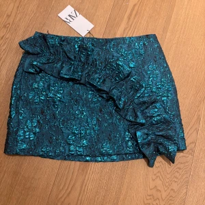 Turkos kjol med volang från Zara - Snygg turkos kjol från Zara med en iögonfallande volangdetalj. Kjolen har en glittrig yta som ger en festlig känsla. Perfekt för att sticka ut med stil.