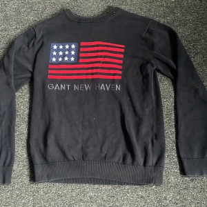 Svart tröja från GANT med flaggmotiv - Snygg svart tröja från GANT med ett stort flaggmotiv i rött, vitt och blått på ryggen. Texten 'GANT NEW HAVEN' är tryckt under flaggan. Perfekt för en stilren och avslappnad look.