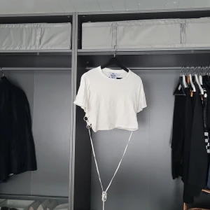 Vit croppad t-shirt från The Ragged Priest - Säljer en vit croppad t-shirt från The Ragged Priest. T-shirten har korta ärmar och en unik detalj med ringar och knytning på sidan. Perfekt för en trendig look! Små hål på ryggen som jag inte sett förrän jag granskade den nu - men jag tänker att det matchar looken :) 