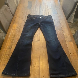 Mörkblå bootcut jeans från True Religion stl. 26 - Snygga mörkblå bootcut jeans från True Religion med klassiska detaljer och kontrastsömmar. De har en låg midja och ikoniska bakfickor med broderi. Perfekta för en avslappnad stil med en touch av retro. Storlek 26 och använda max 5 gånger. Det är dock en liten fläck på en bakfickan, men den syns inte så bra när man har på sig byxorna. Se bild. Jag är 167cm lång och de är precis att det inte släpper i baken.