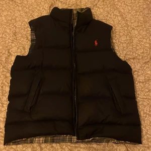 Blå dun väst med rutig insida. Priset kan diskuteras inom rimligt pris - Snygg svart dunväst från Ralph Lauren med en rutig insida i gröna och blå nyanser. Västen har en dragkedja framtill och en broderad logotyp på bröstet. Perfekt för kyligare dagar.