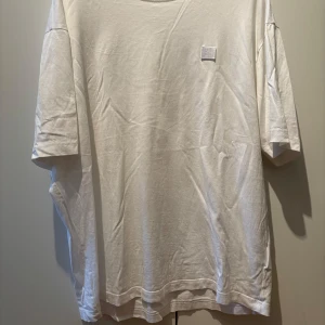 Vit t-shirt från Acne Studios - Säljer en stilren vit t-shirt från Acne Studios i 100% ekologisk bomull. T-shirten har en liten fyrkantig detalj på bröstet och är kortärmad med en normal passform. Perfekt för en minimalistisk look.