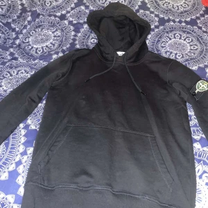 Svart hoodie från Stone Island - Säljer en stilren svart hoodie från Stone Island med klassisk logga på ärmen. Tröjan har en bekväm passform med känguruficka och justerbar huva. Perfekt för en avslappnad look. Pris sänkning vid snabb affär.Den är äkta 