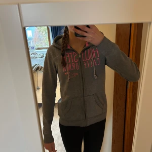 Grå hoodie från Hollister - Säljer en grå luvtröja från Hollister med rosa tryck på bröstet. Tröjan har dragkedja och två fickor framtill. Perfekt för en avslappnad stil. Storlek M men har använt den när jag va mindre så passar Xs-S. Kan sänka priset vid snabb affär💗