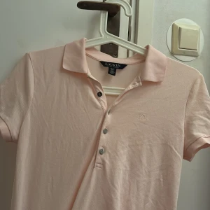 Ljusrosa pikétröja från Ralph Lauren - Säljer en stilren ljusrosa pikétröja från Ralph Lauren. Tröjan har korta ärmar och en klassisk krage med knappar framtill. Perfekt för en avslappnad och snygg look köpt från Ralp louren i somras pris kan diskuteras!.