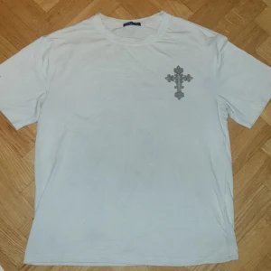 Vit t-shirt med kors och vingar - Säljer en vit t-shirt med ett stort tryck av ett kors och vingar på baksidan. Framsidan har ett mindre kors på bröstet. Perfekt för en unik och stilren look.