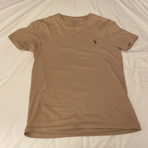 t-shirt Ralph Lauren - Säljer en stilren beige/brun t-shirt från Ralph Lauren med en liten grön broderad logga på bröstet. T-shirten har en slim passform och är kortärmad, perfekt för en sommardag och under en hoodie.