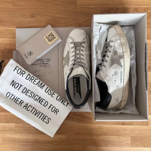 Goldengoose Superstars - Vita Golden Goose Superstars med mörkblå hälkappa. Riktigt bra skick, enda felen är lite bortskrapat i skon på sulan (bild1), liten spricka på innersidan (bild3), och lite hälskrap (bild5). Ingen av felen märks vid användning. Nypris ~5000kr. Allt original medföljer, äkthetbevis finns. 