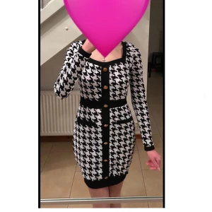 Svartvit mönstrad klänning med knappar - Snygg svartvit klänning med ett klassiskt rutigt mönster och dekorativa knappar framtill. Klänningen har långa ärmar och en figurnära passform som framhäver silhuetten. Perfekt för en stilren look.