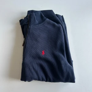 Mörkblå tröja med dragkedja från Ralph Lauren - Säljer en stilren mörkblå tröja från Ralph Lauren med halv dragkedja och röd broderad logotyp på bröstet. Perfekt för en avslappnad men snygg look. Tröjan har långa ärmar och är tillverkad i ett mjukt material.