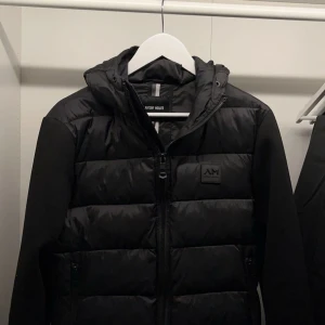 Svart pufferjacka från Antony Morato - Snygg svart pufferjacka från Antony Morato med huva och dragkedja. Jackan har en quiltad design och är perfekt för kyligare dagar. Den har långa ärmar och en stilren look.