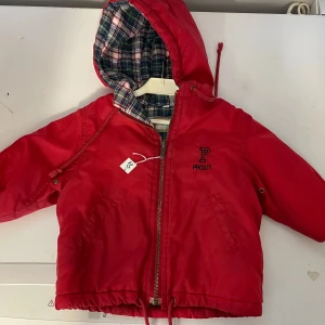 Röd jacka med huva från Prosit - Säljer en röd baby jacka storlek 70 med huva från Prosit. Jackan har ett rutigt foder och dragkedja framtill. Perfekt för kyligare dagar med sin långa ärmar och praktiska design.