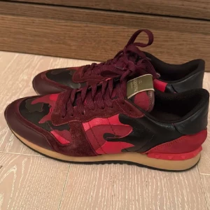 Valentino Rockrunner i röd och svart - Snygga Valentino Rockrunner sneakers i en cool röd och svart camouflage-design. Skorna har snörning och är tillverkade i en kombination av mocka och skinn. Perfekta för att ge din outfit en unik touch.