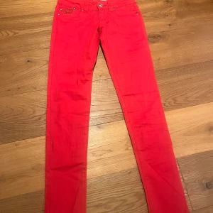 Röda jeansbyxor från Reals - Snygga röda jeansbyxor från Reals med broderad krona på bakfickorna. Byxorna har en slim passform och är dekorerade med nitar vid fickorna. Perfekta för att sticka ut med en djärv färg och coola detaljer.