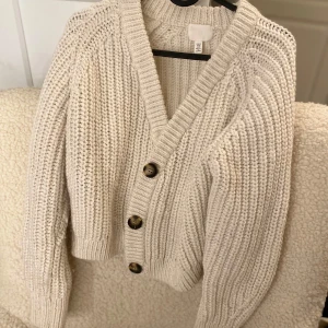 Beige stickad kofta från H&M - Mysig beige stickad kofta från H&M med stora knappar framtill. Perfekt för kyliga dagar med sin ribbade struktur och långa ärmar. En klassisk och stilren design som passar till det mesta.