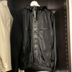 Svart vindjacka från J.Lindeberg - Snygg svart vindjacka från J.Lindeberg med dragkedja framtill och justerbar huva. Jackan har flera praktiska fickor med dragkedjor och en stilren design. Perfekt för blåsiga dagar!