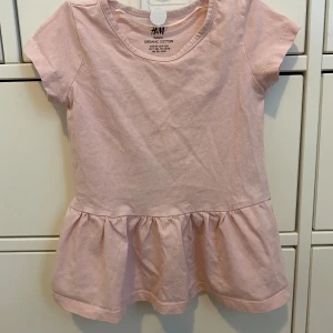 Rosa klänning från H&M - Söt rosa klänning från H&M i ekologisk bomull. Den har korta ärmar och en volangdetalj vid midjan som ger en lekfull look. Perfekt för små fashionistas!