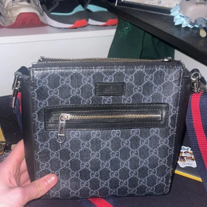 Svart axelväska från Gucci - Snygg svart axelväska från Gucci med det klassiska GG-mönstret. Väskan har en justerbar axelrem i rött och blått, samt en praktisk dragkedja framtill. Perfekt för att bära dina nödvändigheter med stil.