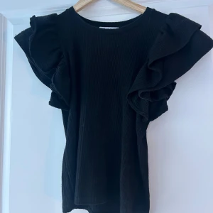 Svart ribbad topp från Zara - Snygg svart ribbad topp från Zara med volangdetaljer på ärmarna. Perfekt för att ge en stilfull touch till din outfit. Toppen har en rund halsringning och korta ärmar med volanger.