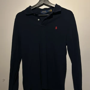 Mörkblå pikétröja från Ralph Lauren - Säljer en klassisk mörkblå pikétröja från Ralph Lauren med den ikoniska röda loggan på bröstet. Tröjan har långa ärmar och en stilren krage med knappar. Perfekt för en avslappnad men stilfull look. Storleken är 14-16 motsvarar xs 