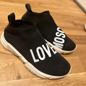 Svarta sneakers från Moschino - Snygga svarta sneakers från Moschino med vit text 'LOVE MOSCHINO' på ovansidan. Skorna har en elastisk stickad överdel och en vit sula. Storlek 39. Köpta för ca 2000:-