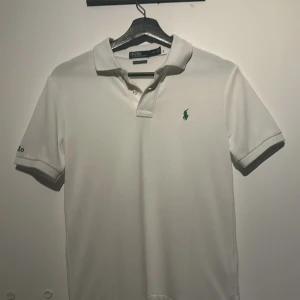 Vit pikétröja från Ralph Lauren - Säljer en klassisk vit pikétröja från Ralph Lauren med den ikoniska gröna loggan på bröstet. Tröjan har en krage och knappar framtill, perfekt för en stilren look. Passar både till vardags och mer uppklädda tillfällen.