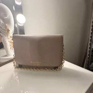 Beige axelväska från Burberry - Snygg beige axelväska från Burberry med klassiskt rutigt mönster och guldfärgad kedja. Väskan har en elegant design med en praktisk insida och justerbar axelrem. Perfekt för att ge din outfit en lyxig touch.