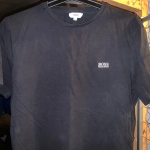 Svart t-shirt från Boss - Säljer en stilren svart t-shirt från Boss med korta ärmar och rund hals. T-shirten har en liten vit logotyp på bröstet. Perfekt för en enkel och snygg look. Barnstorlek 14-16år/ motsvarande S i vuxen